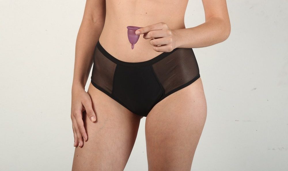 Modelo com calcinha absorvente preta segurando coletor menstrual roxo.