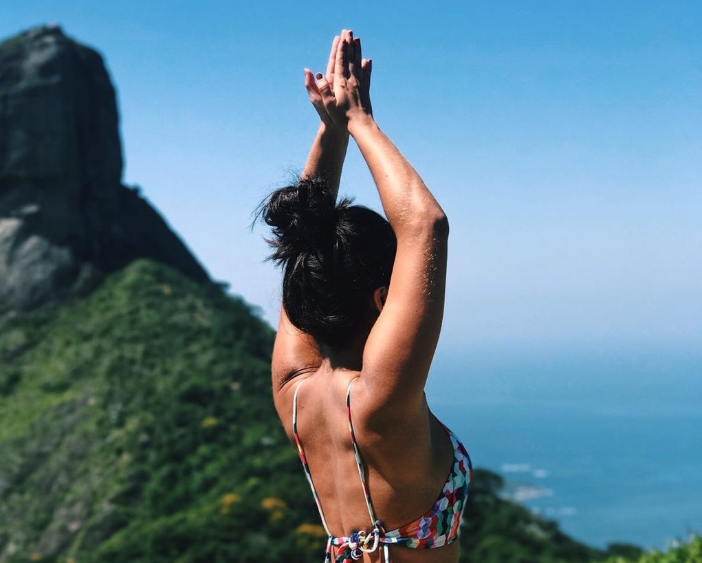 Camila Moll faz ioga no alto de um morro, com o cume ao fundo, o mar e o céu.