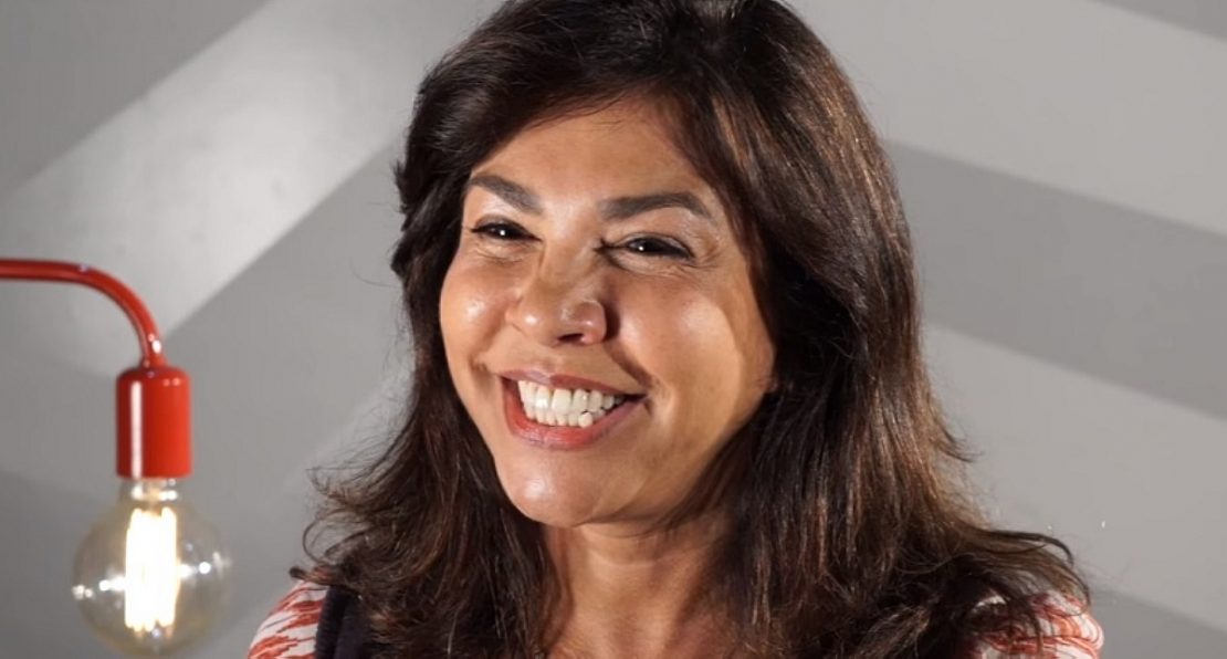 Imagem mostra Mônica Sousa sorrindo. Ela tem cabelos pretos e veste blusa estampada de branco com laranja